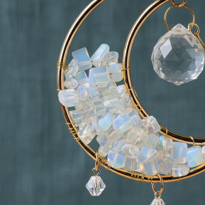 Close-up of a crystal moon suncatcher showing gold crescent frame, iridescent stone chips, and a faceted prism crystal against a teal linen background. — for Carpe Diem Dream