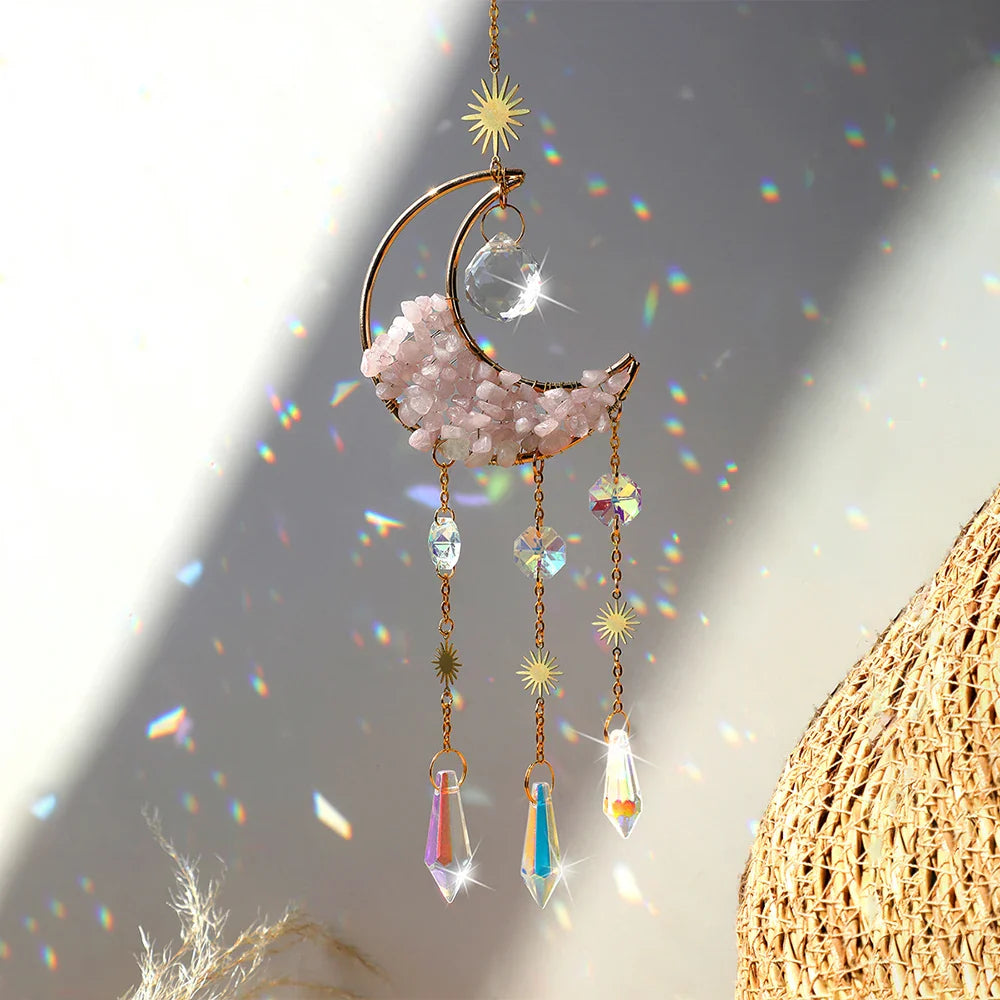 A rose quartz crystal moon suncatcher with a gold crescent frame, soft pink stone chips, and hanging prism drops displayed in warm natural sunlight with sparkling rainbow reflections and gentle shadows. — for Carpe Diem Dream