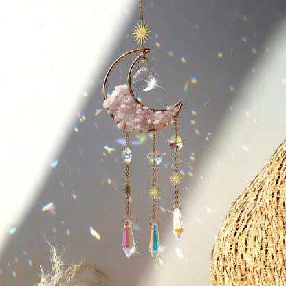 A rose quartz crystal moon suncatcher with a gold crescent frame, soft pink stone chips, and hanging prism drops displayed in warm natural sunlight with sparkling rainbow reflections and gentle shadows. — for Carpe Diem Dream