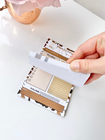 Hand lifting a sheet from a minimalist 4x6 daily planning sticky notepad with priority, checklist, water intake, and gratitude sections on a white desk surface. — for Carpe Diem Dream