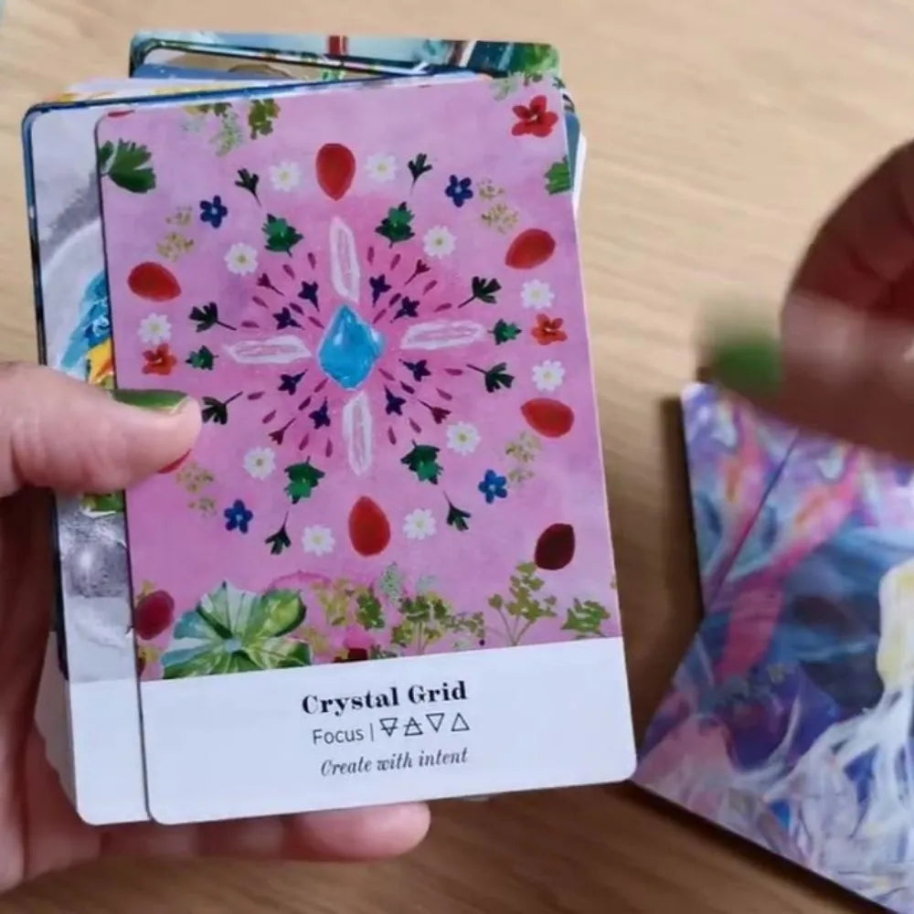 A hand holds an Earth Alchemy Oracle card with a crystal grid design while the rest of the deck is visible in the background on a wooden surface. — for Carpe Diem Dream
