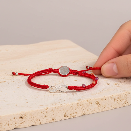 Red string bracelet with a silver infinity charm shown in true scale on a travertine surface as a hand adjusts the cord under soft natural lighting. — for Carpe Diem Dream