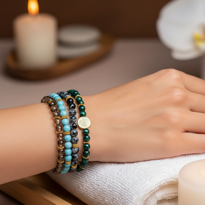 A stacked set of four crystal bracelets featuring turquoise, malachite, smoky quartz, and snowflake obsidian worn on the wrist in a serene spa setting with a folded towel and candle. — for Carpe Diem Dream