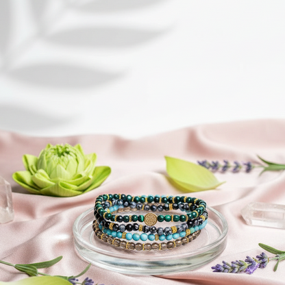 A stacked set of four crystal bracelets featuring turquoise, malachite, smoky quartz, and snowflake obsidian displayed on a glass dish with soft botanicals and crystals on a pale pink fabric backdrop. — for Carpe Diem Dream