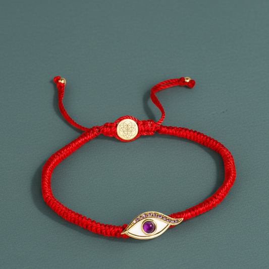 A red string protection bracelet with an amethyst Evil Eye charm and gold-plated details displayed on a muted teal surface. — for Carpe Diem Dream