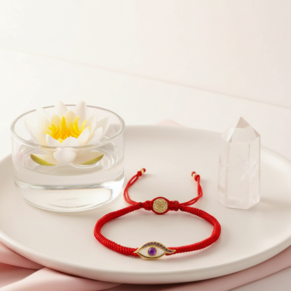 A red string protection bracelet with an amethyst Evil Eye charm arranged on a round white tray beside a floating lotus and a clear quartz point in soft natural light. — for Carpe Diem Dream
