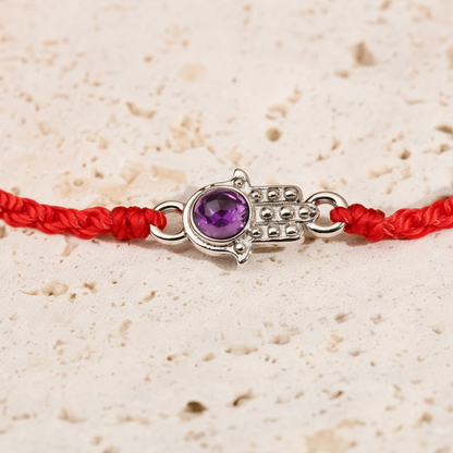 Close-up view of the silver Hamsa charm with amethyst on a red string bracelet displayed on a textured travertine surface. — for Carpe Diem Dream