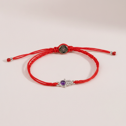 Red string bracelet with a silver Hamsa charm and amethyst displayed on softly lit blush silk fabric. — for Carpe Diem Dream