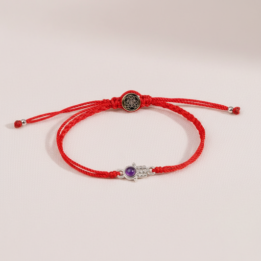 Red string bracelet with a silver Hamsa charm and amethyst displayed on softly lit blush silk fabric. — for Carpe Diem Dream