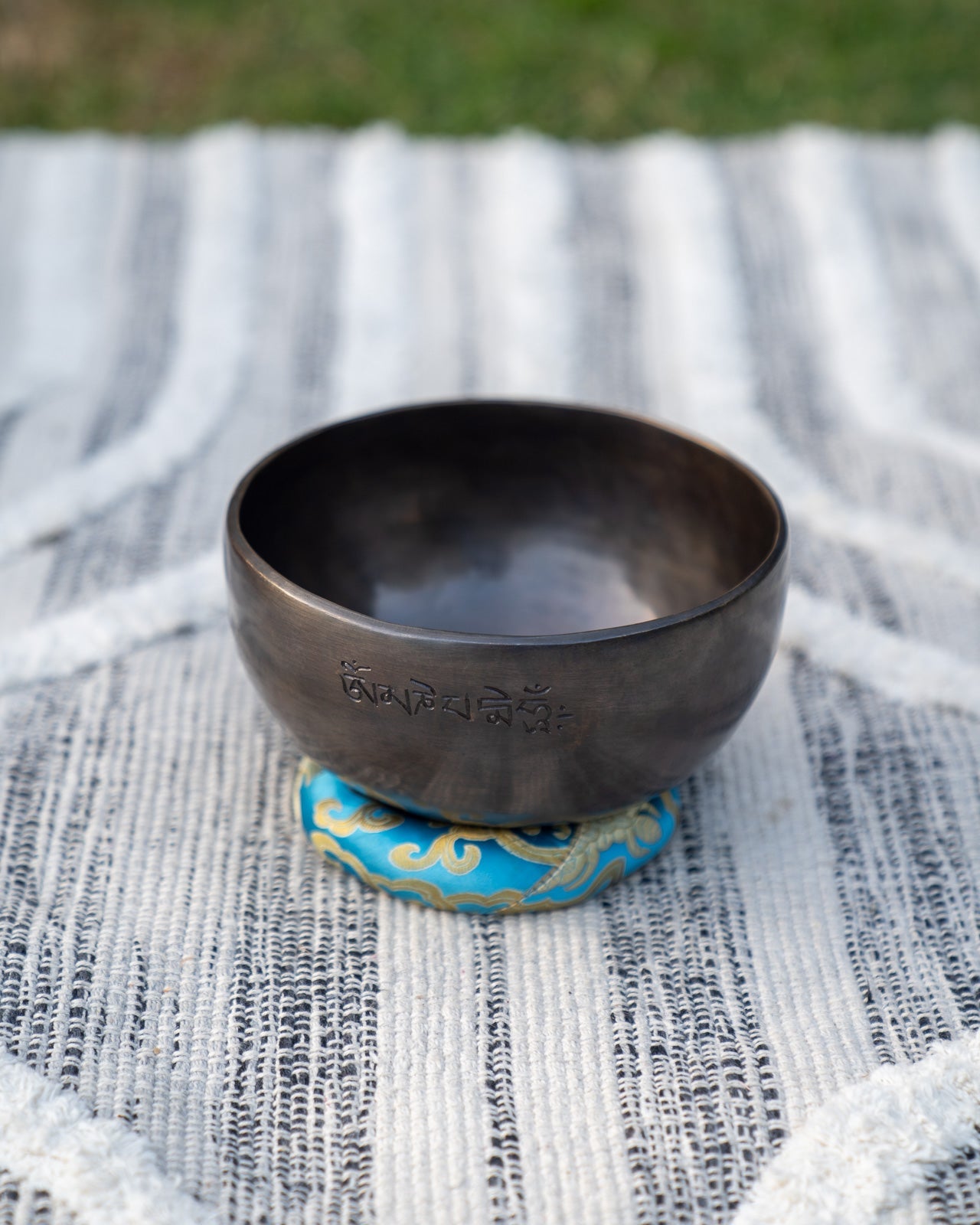Back view of a Tibetan singing bowl with engraved mantra resting on a blue embroidered cushion outdoors. — for Carpe Diem Dream