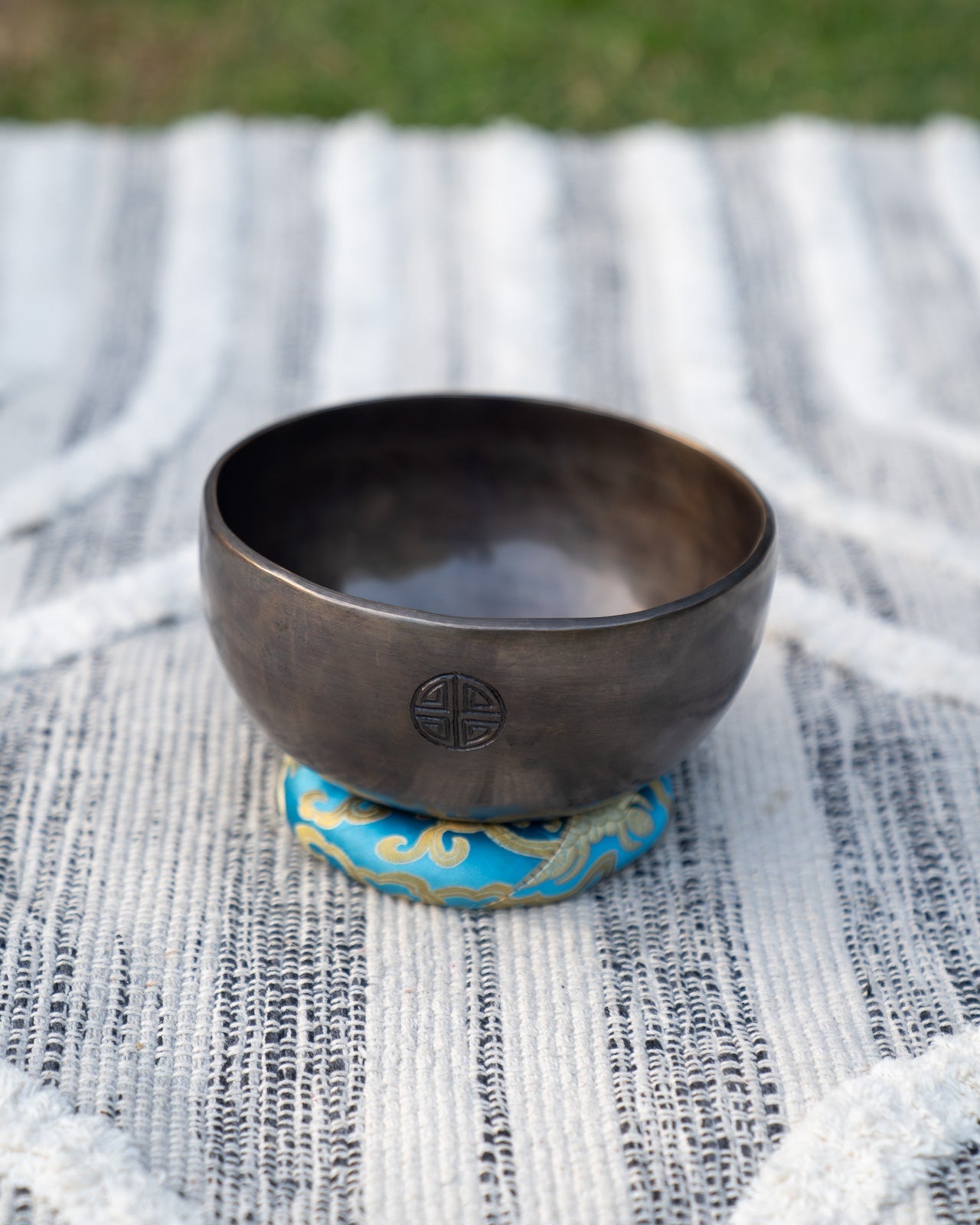 Throat chakra Tibetan singing bowl with engraved full moon symbol resting on a blue embroidered cushion outdoors on a natural surface. — for Carpe Diem Dream