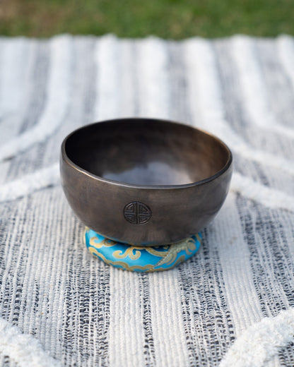 Throat chakra Tibetan singing bowl with engraved full moon symbol resting on a blue embroidered cushion outdoors on a natural surface. — for Carpe Diem Dream