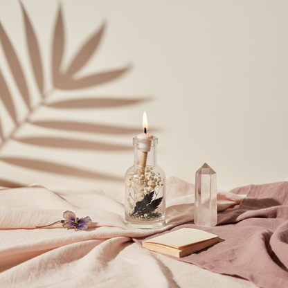 A Gemini zodiac oil candle in a clear glass vessel with embedded botanicals is styled on soft draped fabric beside a crystal point and a small book with botanical shadows on a light neutral wall. — for Carpe Diem Dream