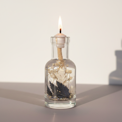 A Gemini zodiac oil candle in a clear glass vessel with embedded botanicals burns with a steady flame on a clean neutral surface with soft natural shadows. — for Carpe Diem Dream