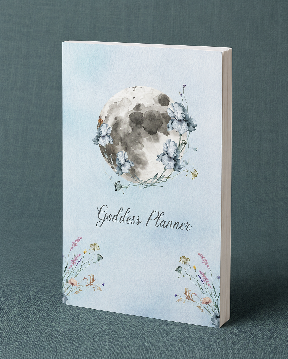 Goddess Planner paperback displayed upright on a teal-grey linen background featuring watercolor moon and floral artwork, designed for lunar planning, self-care reflection, habit tracking, and intentional living. — for Carpe Diem Dream
