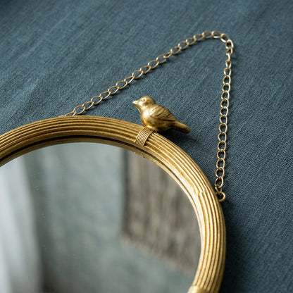 Close-up of a gold bird perched atop a ribbed gold wall mirror frame, showing the decorative hanging chain against a cool blue-gray textured background.
