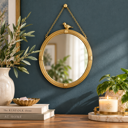 Round gold hanging wall mirror with decorative bird accent and gold chain displayed on a dark teal wall above a wooden console with botanical accents, books, and a candle.