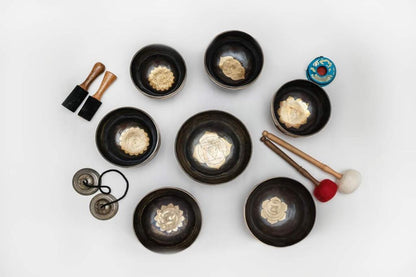 Seven gold mandala Tibetan singing bowls with embroidered chakra cushions, wooden mallets, rotating sticks, and tingsha cymbals displayed together on a white background. — for Carpe Diem Dream