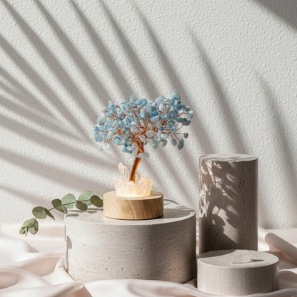 Aquamarine crystal angel tree lamp glowing softly on stone pedestals with eucalyptus accents and dramatic shadow patterns against a textured white backdrop. — for Carpe Diem Dream