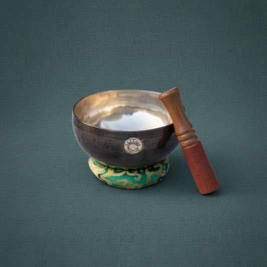 A hand-hammered Heart Chakra singing bowl with wooden striker resting on a green cushion against a muted teal linen background. — for Carpe Diem Dream