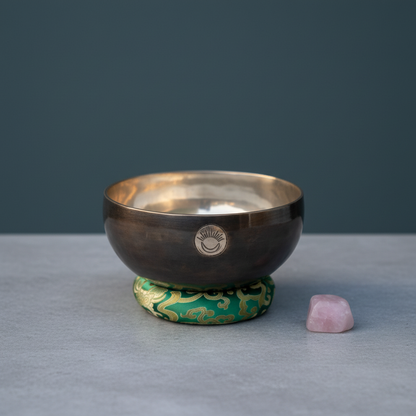 A Heart Chakra singing bowl resting on a green cushion beside a rose quartz crystal on a soft neutral surface with a muted blue background. — for Carpe Diem Dream