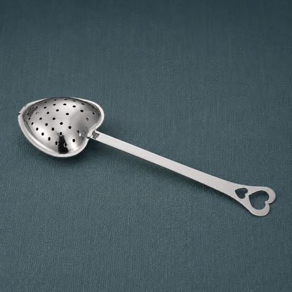 Heart Shaped Tea Infuser – authentic silver heart-shaped metal tea infuser displayed lying flat on a dark teal textured linen backdrop with matching linen surface, photographed in cool diffused daylight with soft natural shadows and premium commercial styling. The product remains the sole focal point with no props. — for Carpe Diem Dream