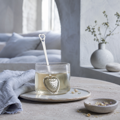 Heart Shaped Tea Infuser – authentic silver heart-shaped metal tea infuser steeping in a clear glass cup of tea within a serene quiet-luxury wellness space with Roman Clay textures, natural stone, soft blue linen, dried herbs, and cool diffused daylight. — for Carpe Diem Dream