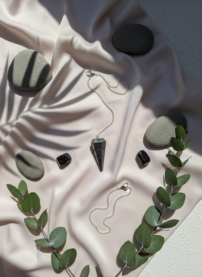 Hematite crystal pendulum resting on soft ivory silk with travertine tiles, grounding stones, and eucalyptus leaves in natural sunlight – for Carpe Diem Dream.