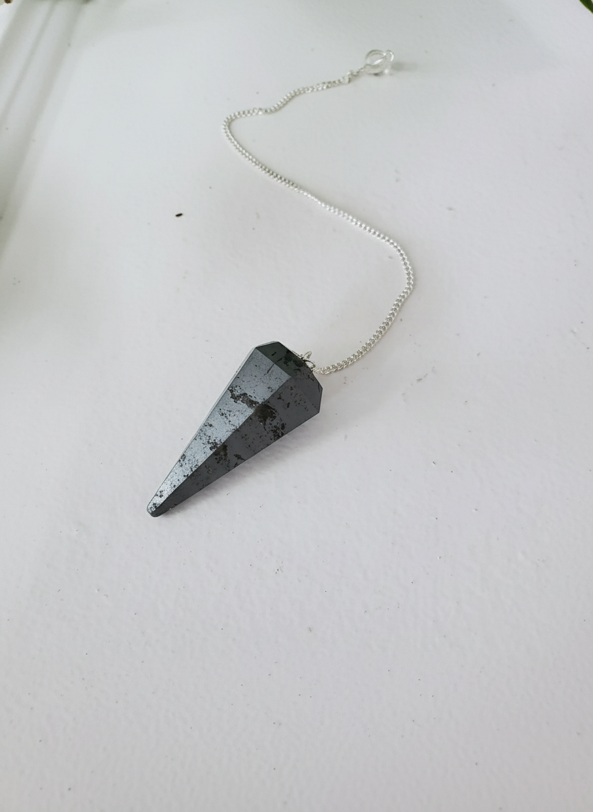 Hematite crystal pendulum with silver chain displayed on a clean white surface, capturing its metallic sheen and grounding energy – for Carpe Diem Dream.
