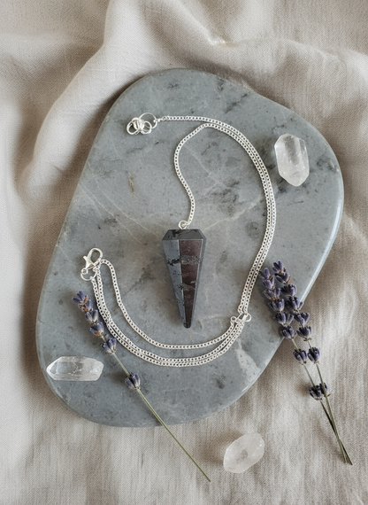 Hematite crystal pendulum displayed on a gray marble tray with lavender sprigs and clear quartz crystals, symbolizing grounding and inner calm – for Carpe Diem Dream.