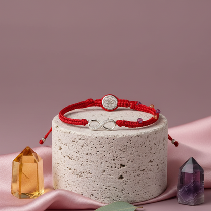 Red string bracelet with a silver infinity charm displayed on smooth travertine pedestals with soft pink draping and gentle natural lighting. — for Carpe Diem Dream