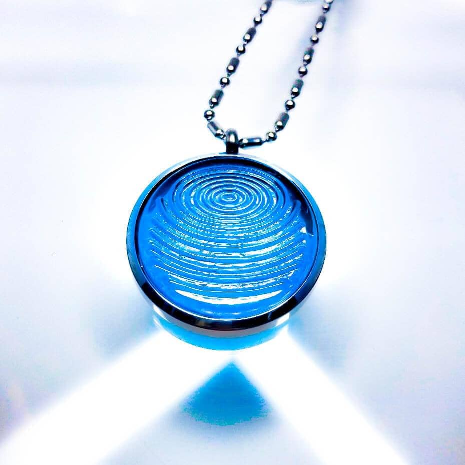 A dark aqua blue ion glass defense pendant with a spiral design on a silver chain, displayed with bright lighting on a white background. — for Carpe Diem Dream