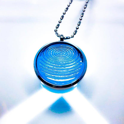 A dark aqua blue ion glass defense pendant with a spiral design on a silver chain, displayed with bright lighting on a white background. — for Carpe Diem Dream