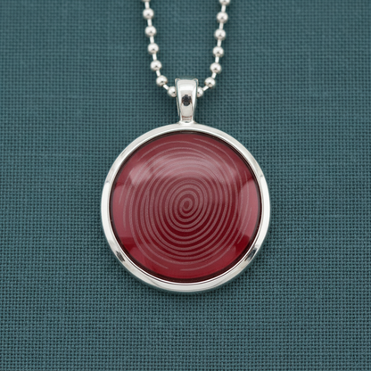 A round ion glass defense pendant with a red spiral design set in a silver frame, displayed on a teal textured surface in soft natural light. — for Carpe Diem Dream