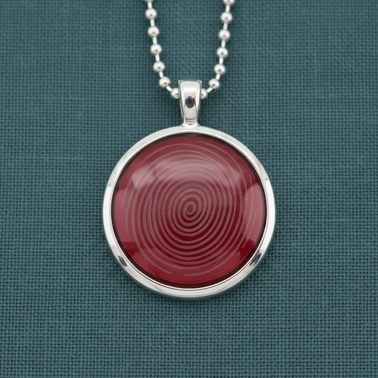 A round ion glass defense pendant with a red spiral design set in a silver frame, displayed on a teal textured surface in soft natural light. — for Carpe Diem Dream