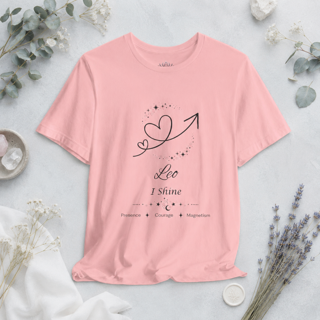 Leo Rising I Shine zodiac T-shirt in pink with black celestial heart and arrow artwork and Presence, Courage, Magnetism text, styled in a cool-toned studio botanical flat lay. — for Carpe Diem Dream