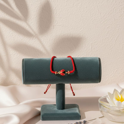 A red string bracelet with a gold-plated ladybug charm displayed on a teal velvet stand beside a floating lotus blossom in soft natural light. — for Carpe Diem Dream