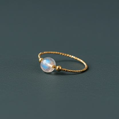 Gold moonstone ring with twisted band and luminous crystal bead on soft light backdrop. — for Carpe Diem Dream