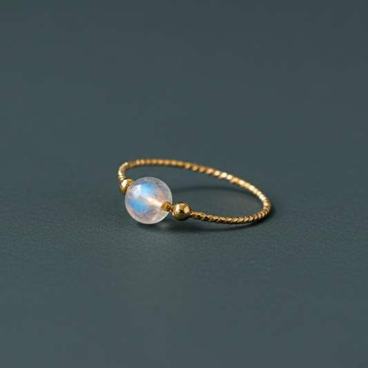 Gold moonstone ring with twisted band and luminous crystal bead on soft light backdrop. — for Carpe Diem Dream
