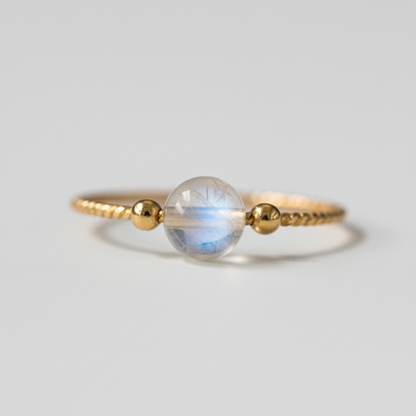 Gold moonstone ring with twisted band and glowing crystal bead on soft neutral backdrop. — for Carpe Diem Dream