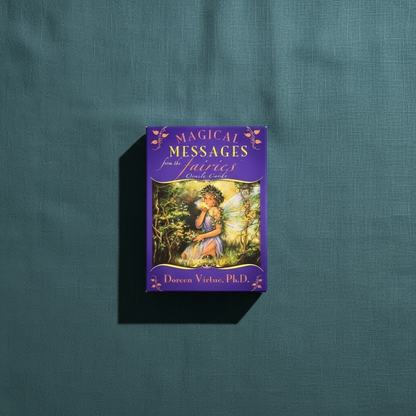 A Magical Messages From The Fairies Oracle deck box with colorful fairy artwork displayed upright on a deep teal textured linen surface with soft directional lighting. — for Carpe Diem Dream