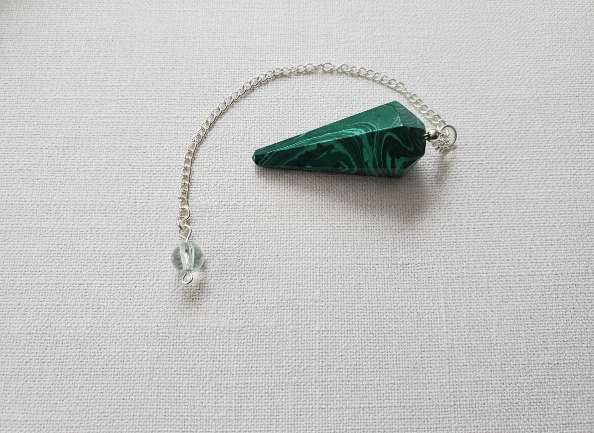Malachite crystal pendulum with silver chain resting on a textured white surface, showcasing deep green swirls and natural patterning – for Carpe Diem Dream.