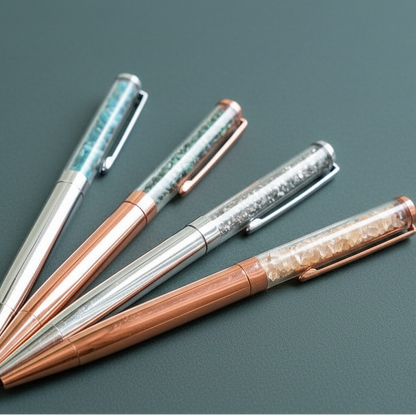 Four crystal-infused pens in silver and rose gold finishes arranged in a flatlay on a muted teal surface, showcasing their Citrine, Malachite, Pyrite, and Apatite gemstone chambers. — for Carpe Diem Dream