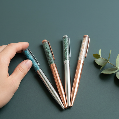 A hand reaches toward four crystal-infused pens resting on a muted teal surface beside a small eucalyptus sprig, showing scale and natural texture. — for Carpe Diem Dream