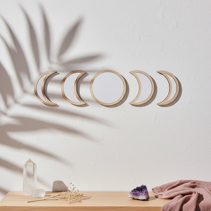Five-piece moon phase wall mirror set with beige wood frames displayed on a textured white wall above a light wood shelf with crystals, an amethyst cluster, dried florals, a mauve draped cloth, and soft botanical shadows. — for Carpe Diem Dream