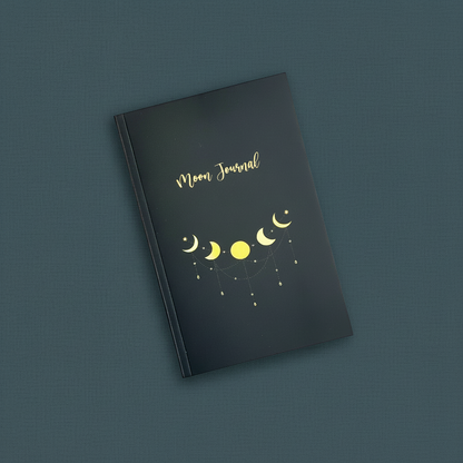 Moon Rituals & Manifestation Journal with black glossy softcover and gold moon phase design displayed upright on a textured teal background — for Carpe Diem Dream