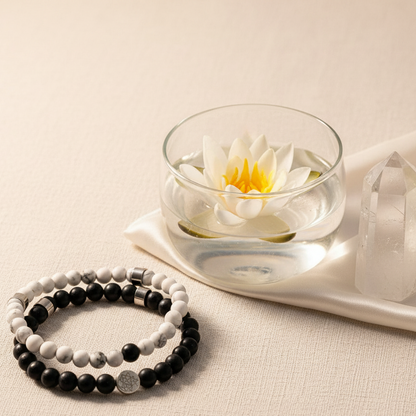 A pair of black and white gemstone bracelets rests on a cream satin surface beside a glass bowl with a floating lotus and a clear quartz point in soft natural light. — for Carpe Diem Dream