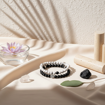 A pair of black and white gemstone bracelets displayed on a cream satin surface beside a glass bowl with a floating lotus and natural shadows. — for Carpe Diem Dream