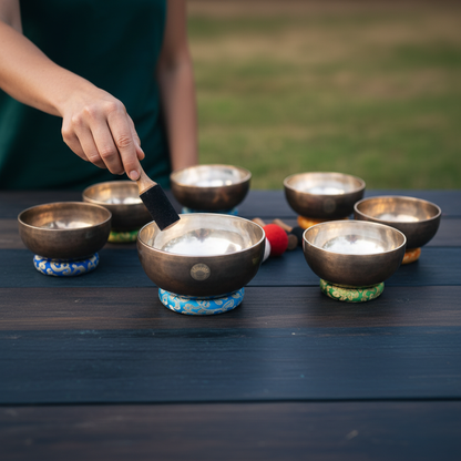 Person striking a Tibetan singing bowl with a wooden mallet while seven chakra singing bowls rest on embroidered cushions on a dark wooden table outdoors. — for Carpe Diem Dream