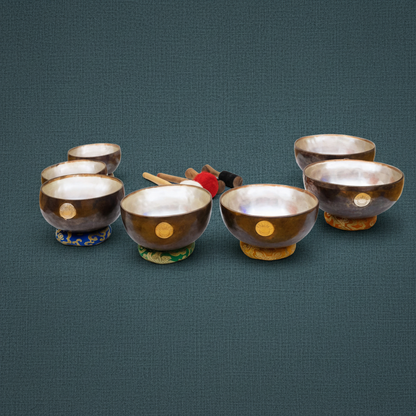 Seven chakra singing bowls with engraved sun and moon emblems resting on embroidered cushions with wooden mallets arranged behind them on a muted teal textured background. — for Carpe Diem Dream